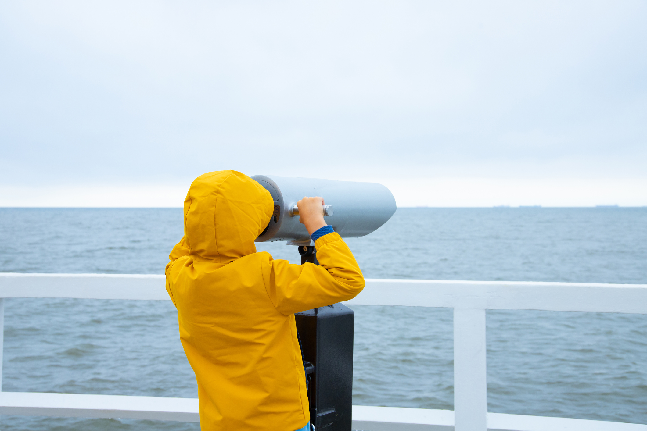 A child in a jacket stands on a white pier. looking through binoculars at the sea. Cold weather. back view